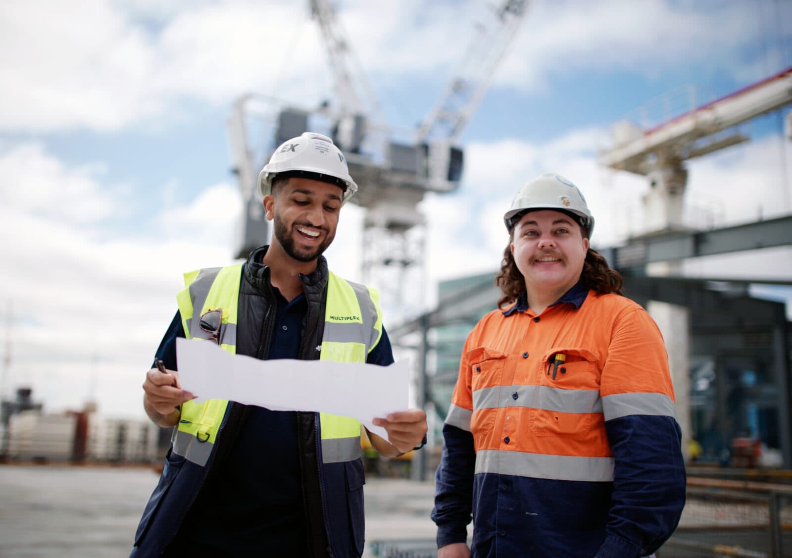 Apprentice and foreman looking at plans with cranes behind them in the distance